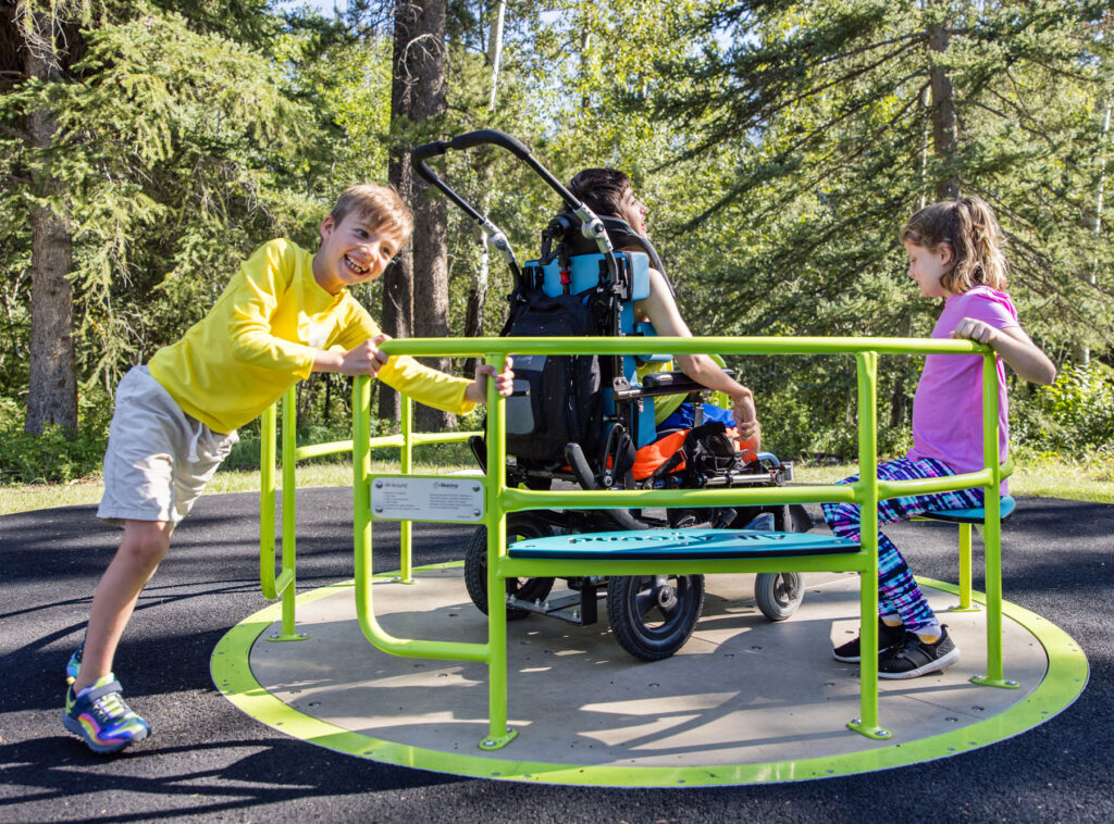 Boy in yellow shirt helps to spin the wheelchair-accessible merry-go-round with two kids in it including one child in a wheelchair