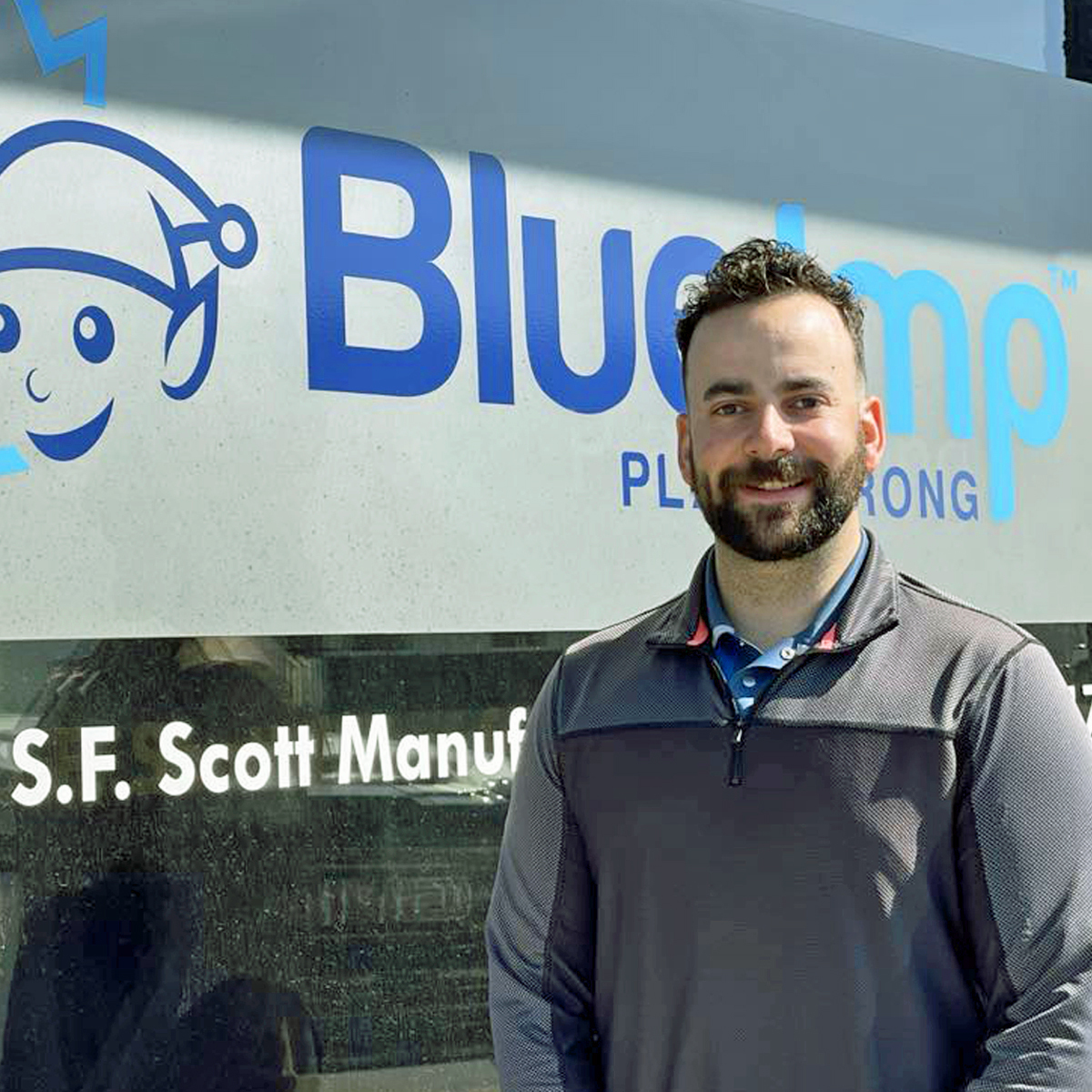 Zach Zubrecki, a playground consultant at Blue Imp head office since 2018, smiling and wearing a casual shirt, surrounded by playground equipment design plans and park furnishings.